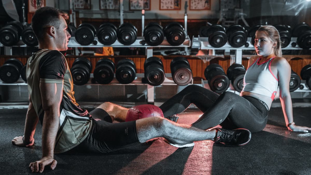 A man and woman resting together on the gym floor, surrounded by dumbbells.